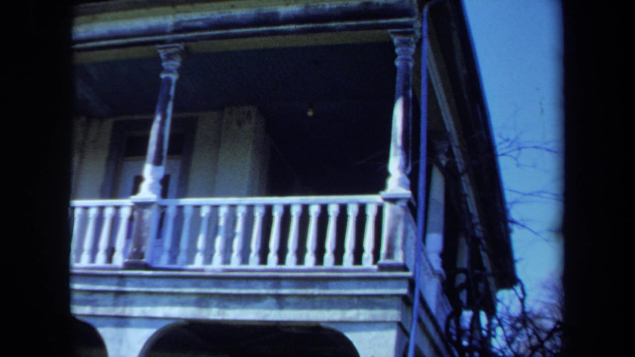 1971 Maine 8mm frame of a white two-story veranda with turned railings, columns, shadowed windows, and bare branches nearby.