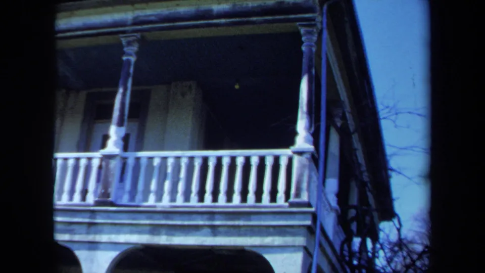 1971 Maine 8mm frame of a white two-story veranda with turned railings, columns, shadowed windows, and bare branches nearby.