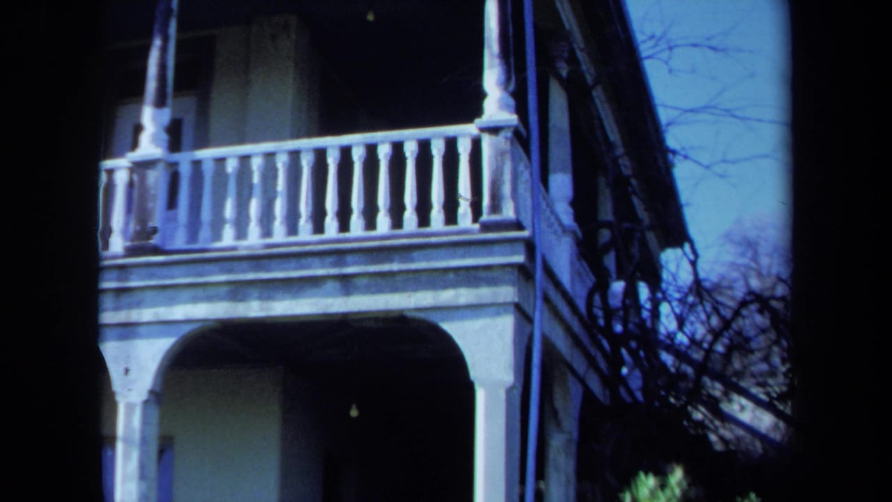 1971 Maine home-movie frame showing the corner of a white two-story porch, upper balustrade, columns, and winter-bare branches.
