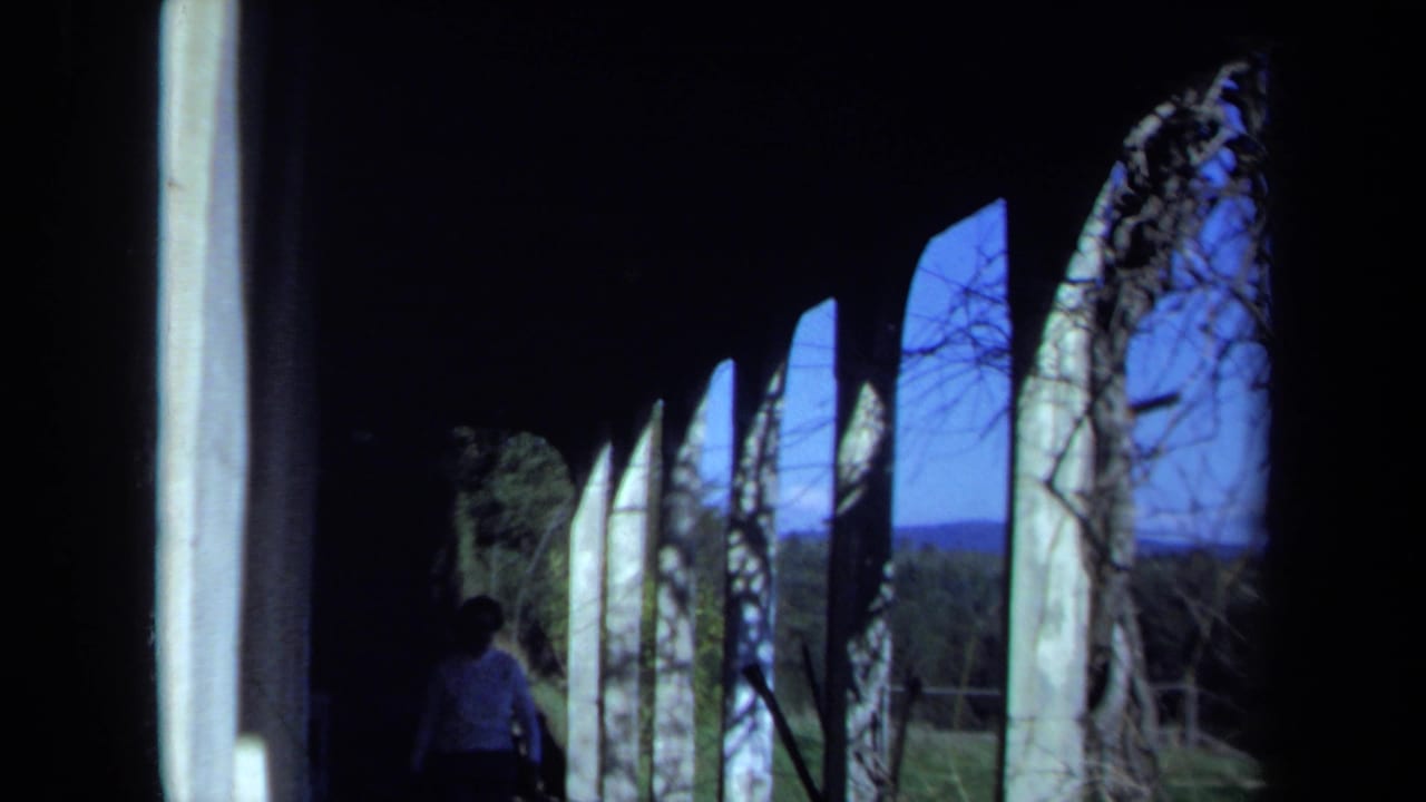 1971 Maine home-movie frame of a covered veranda walkway, arched supports, lawn beyond, and a woman in a light shirt walking through shadow.