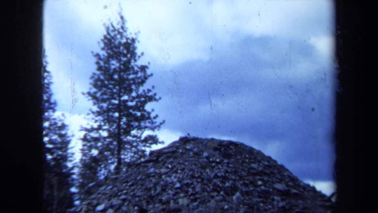 1971 Maine 8mm frame showing a hilltop pile of dark rock or soil with evergreen trees under a cloudy blue-gray sky.