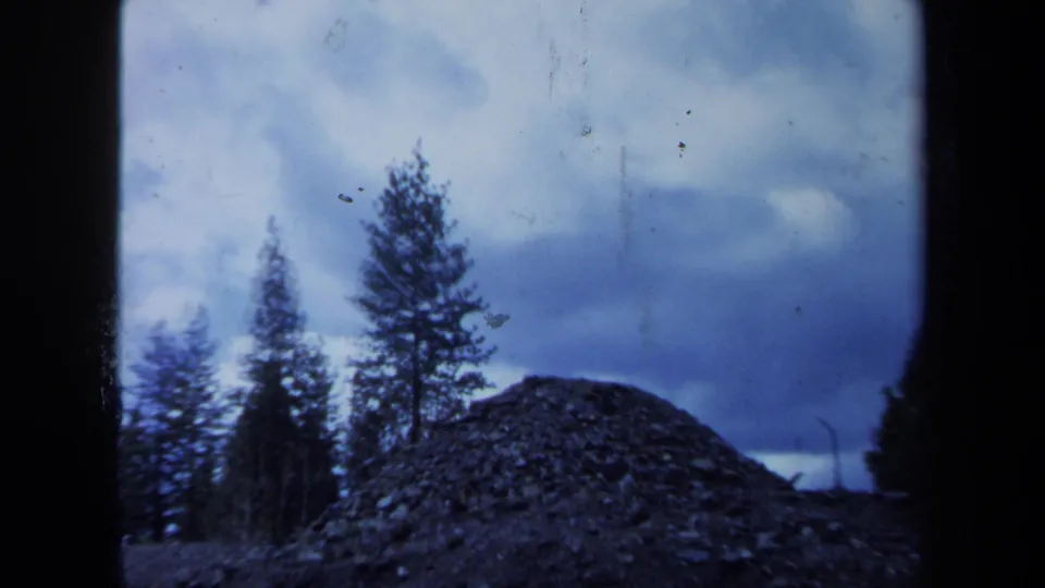 1971 Maine 8mm frame showing a dark mound of stones or earth with evergreen trees and a cloudy sky beyond.