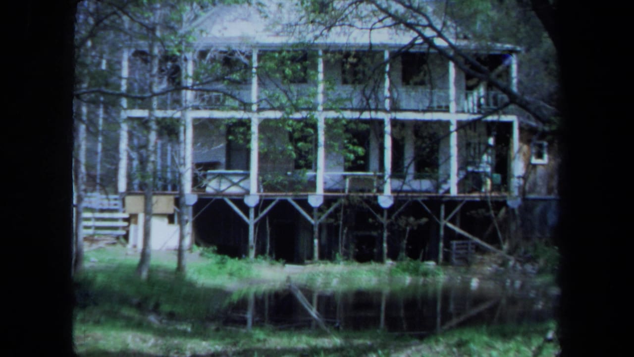 1971 Maine home-movie frame of a large stilted house with broad balconies, surrounded by trees and reflected in water.
