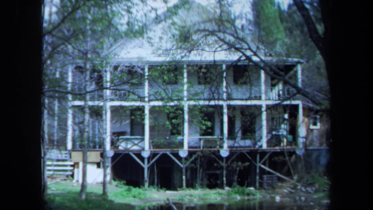 1971 Maine 8mm view of the large house, showing stacked balconies, white railings, trees, and shadowed supports below.