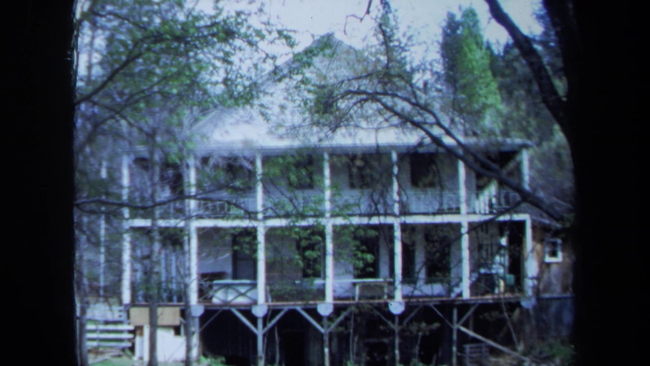 1971 Maine frame of the balcony-fronted house viewed through trees, with grass and a small pond or wet foreground below.