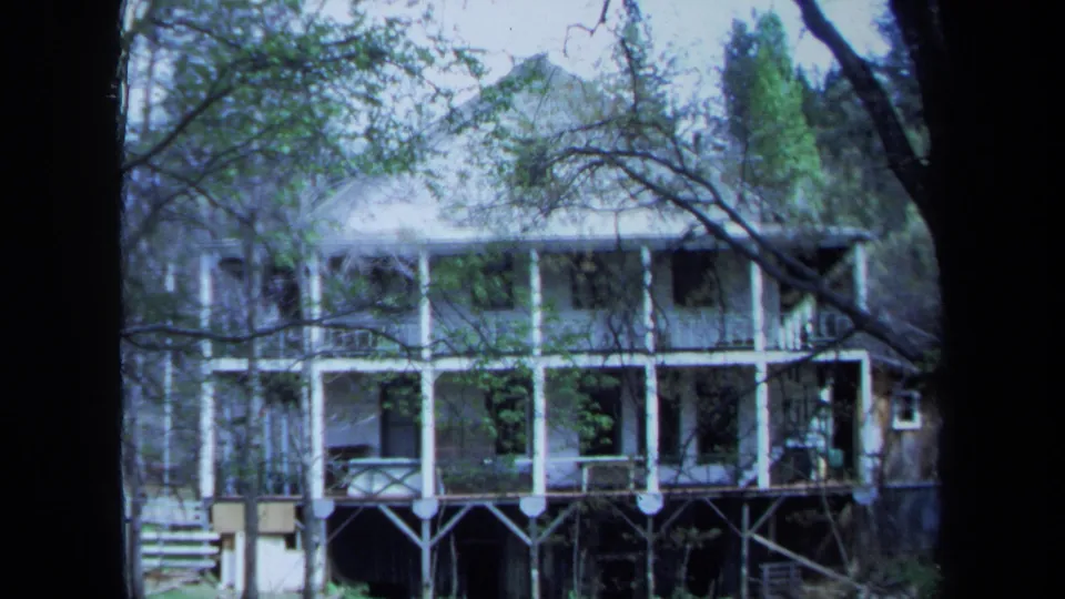 1971 Maine frame of the balcony-fronted house viewed through trees, with grass and a small pond or wet foreground below.