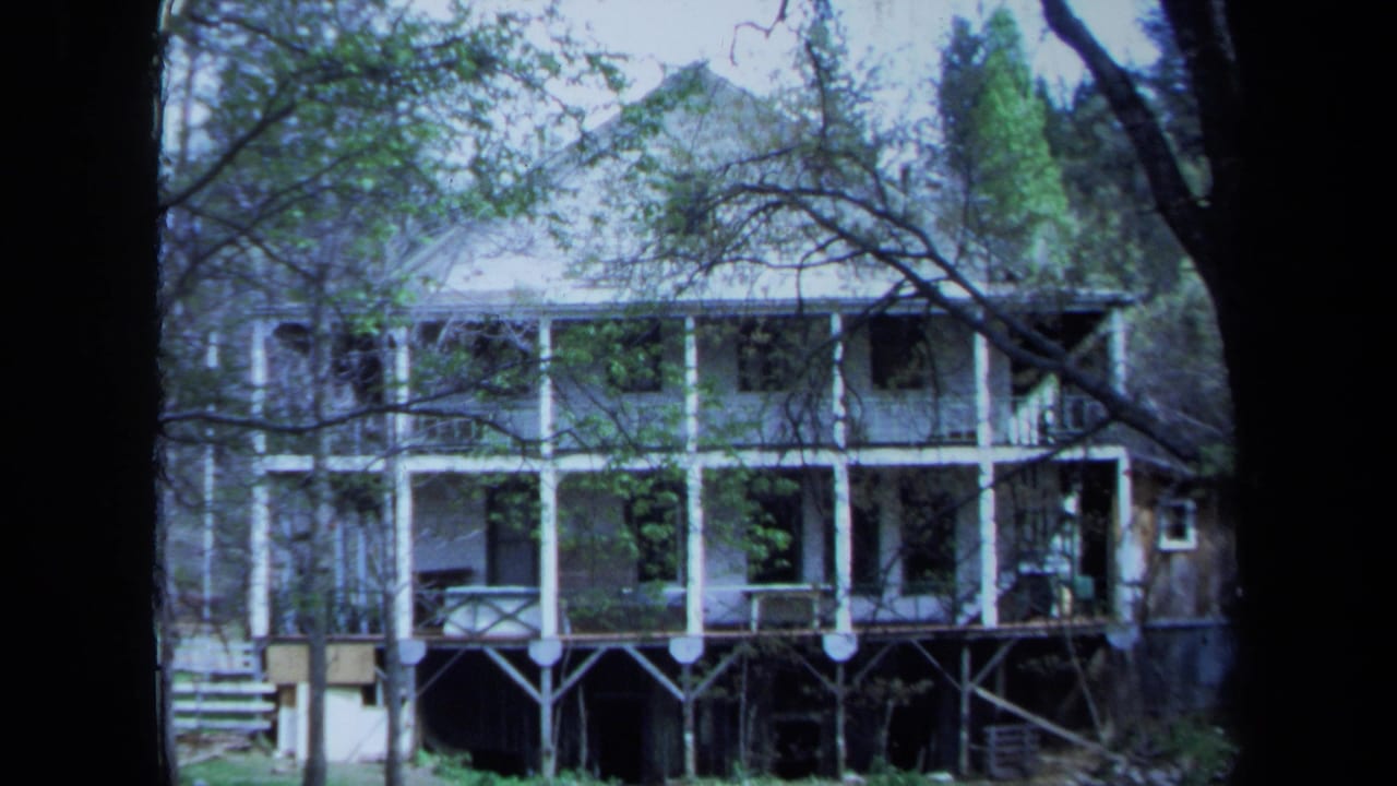 Two-story house with white porches seen through dark trees and branches.