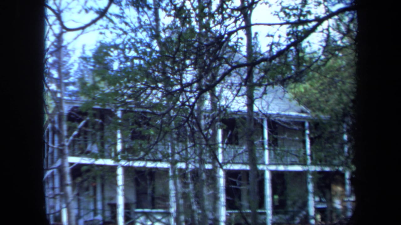 A 1971 Maine 8mm still frames the porch-fronted house through dense branches, with white supports and dark shaded windows visible.