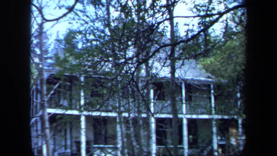 A 1971 Maine 8mm still frames the porch-fronted house through dense branches, with white supports and dark shaded windows visible.