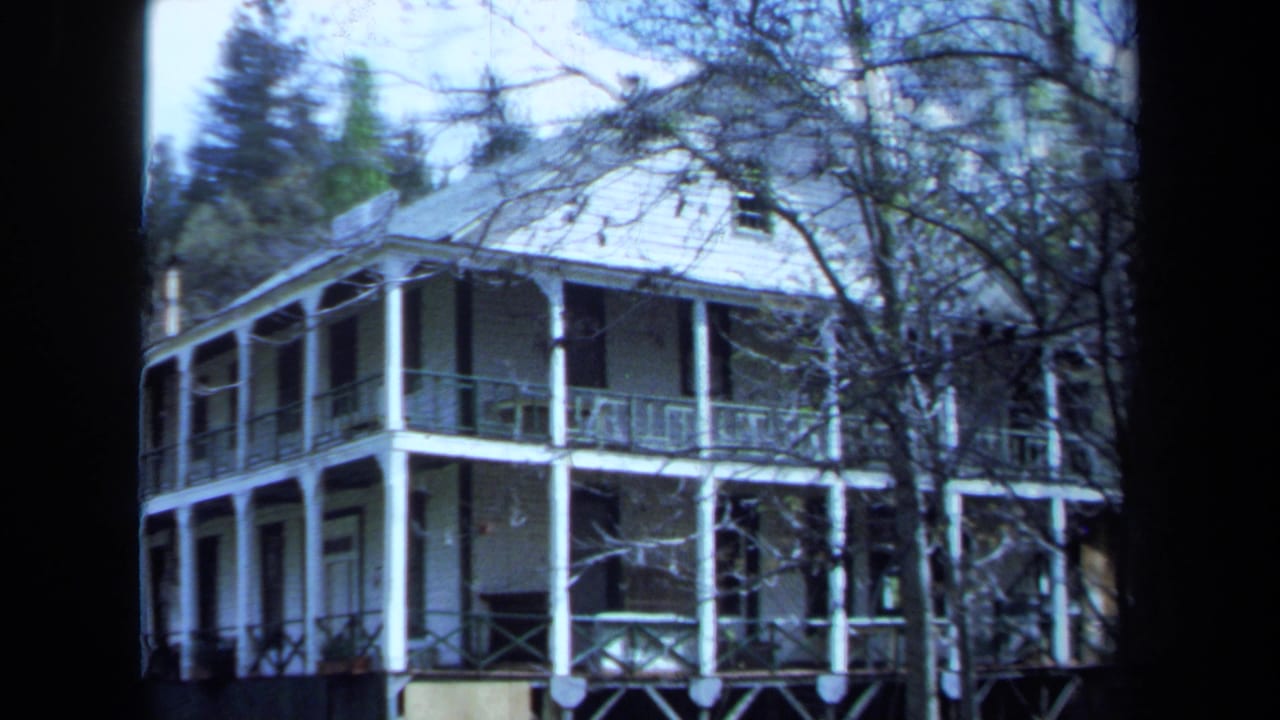 1971 Maine 8mm frame of a large white house with wraparound porches, columns, railings, and a sloped roof among trees.