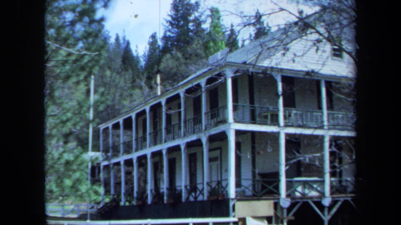 Two-story wooden house with porches among pine trees.