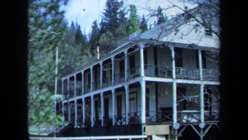 1971 Maine 8mm frame showing a two-story wooden house with white porch columns, dark railings, and surrounding pine trees.