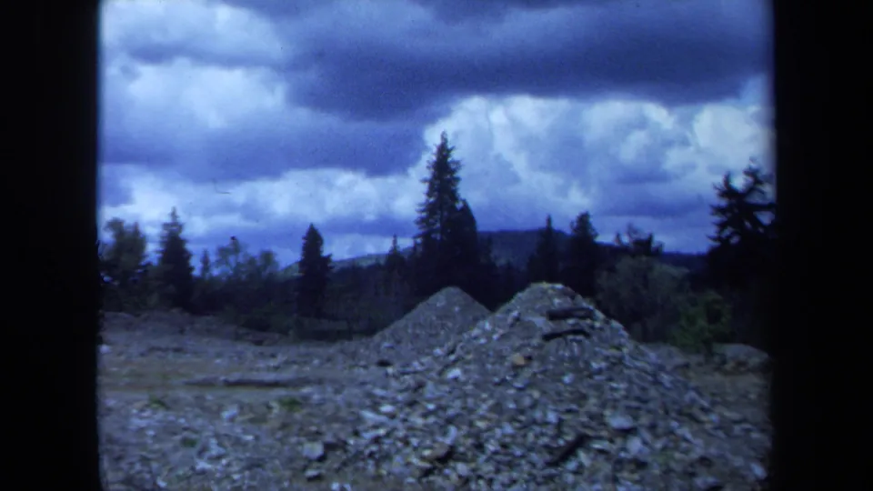 1971 Maine 8mm frame showing gravel piles or rough ground, distant trees, and a heavy overcast sky.