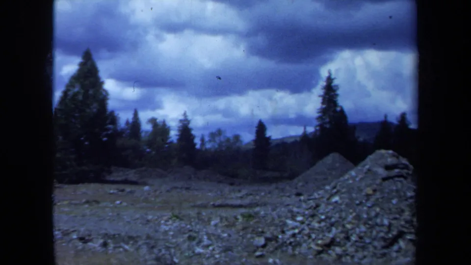 1971 Maine home-movie frame of a gravel mound, open cleared terrain, silhouetted trees, and layered storm clouds.