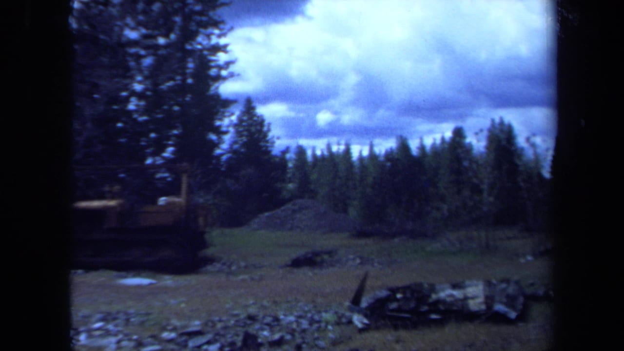 1971 Maine home-movie frame showing a yellow bulldozer beside gravel and debris in an evergreen clearing under a cloudy sky.