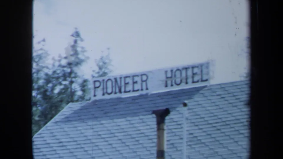 1971 Maine frame holds on the Pioneer Hotel roof sign, shingled roof, chimney pipe, trees, and pale sky.