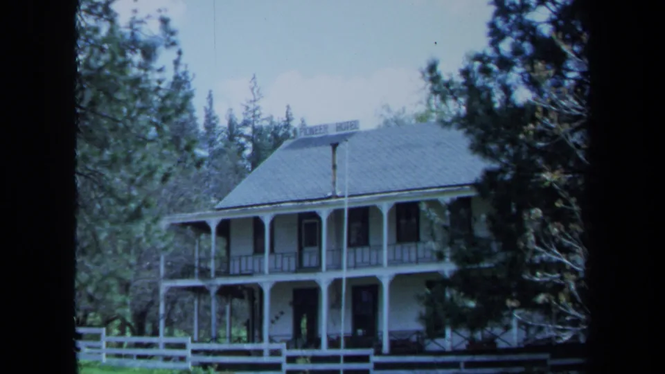 Wider 1971 Maine 8mm view of the white two-story Pioneer Hotel with wraparound porch, fence, roof sign, and surrounding trees.