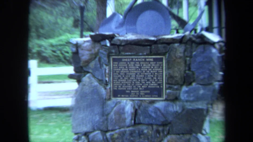 Wider 1971 home-movie view of the Sheep Ranch Mine marker with a white fence, grass, trees, and mining-related metal shapes above the plaque.