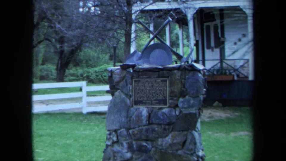 1971 8mm view of the Sheep Ranch Mine marker on a lawn near a white porch-fronted building, with trees and a fence in the background.
