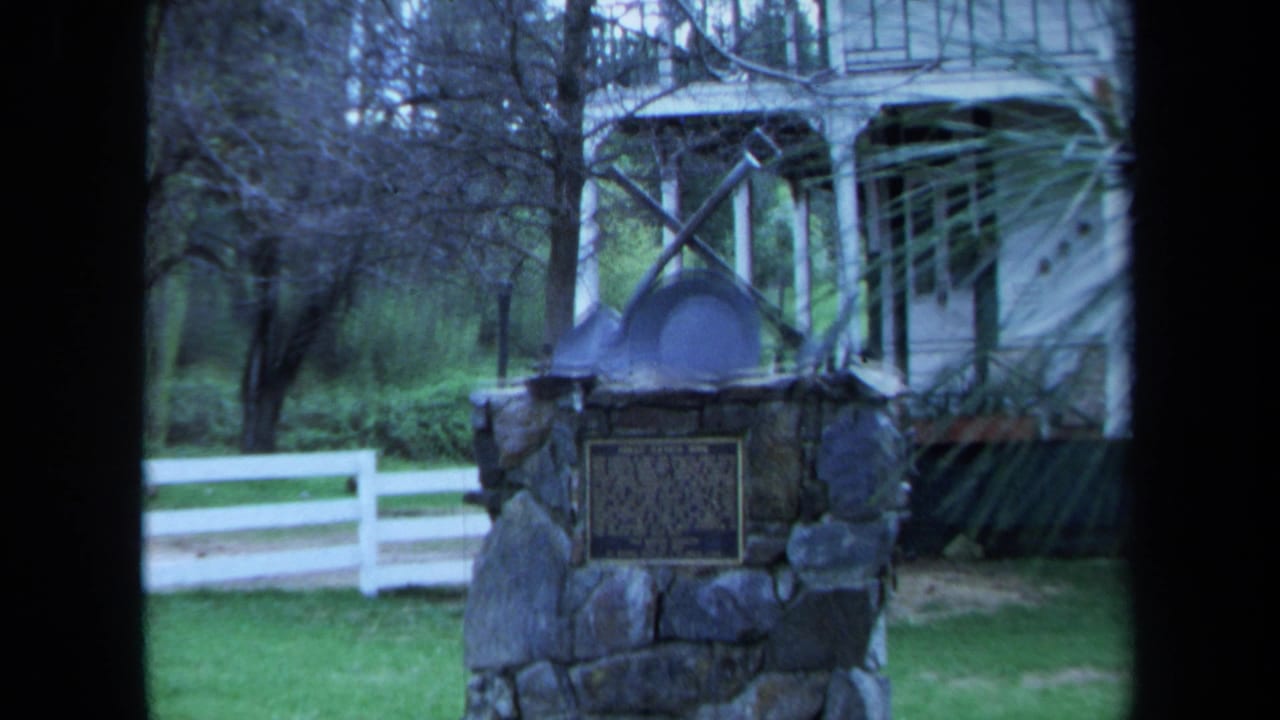 1971 Maine 8mm frame showing a stone marker with plaque, crossed tools, white fence, and part of a farmhouse behind trees.