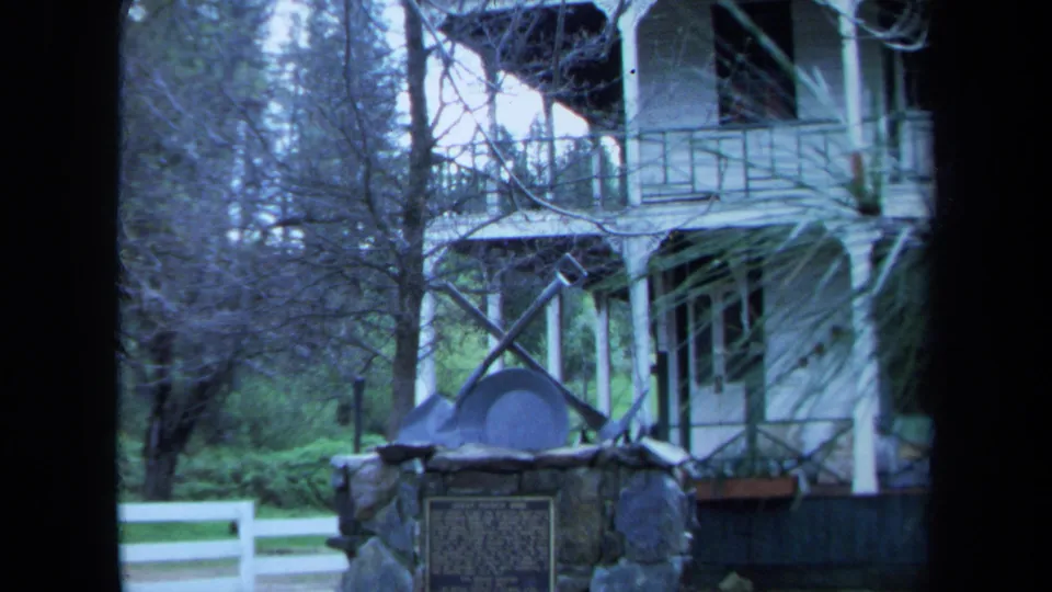 1971 Maine home-movie frame of a white farmhouse, porch, stone marker, and fence partly obscured by dark frame edges and branches.