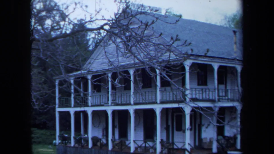 1971 Maine 8mm frame showing a white two-story farmhouse with long porch, dark windows, lawn, and leafless foreground branches.