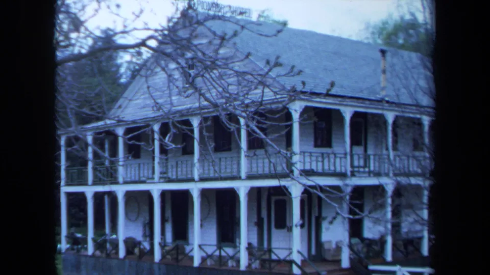 1971 Maine home-movie frame of the farmhouse roofline, porch, railings, lawn, and bare branches across the foreground.