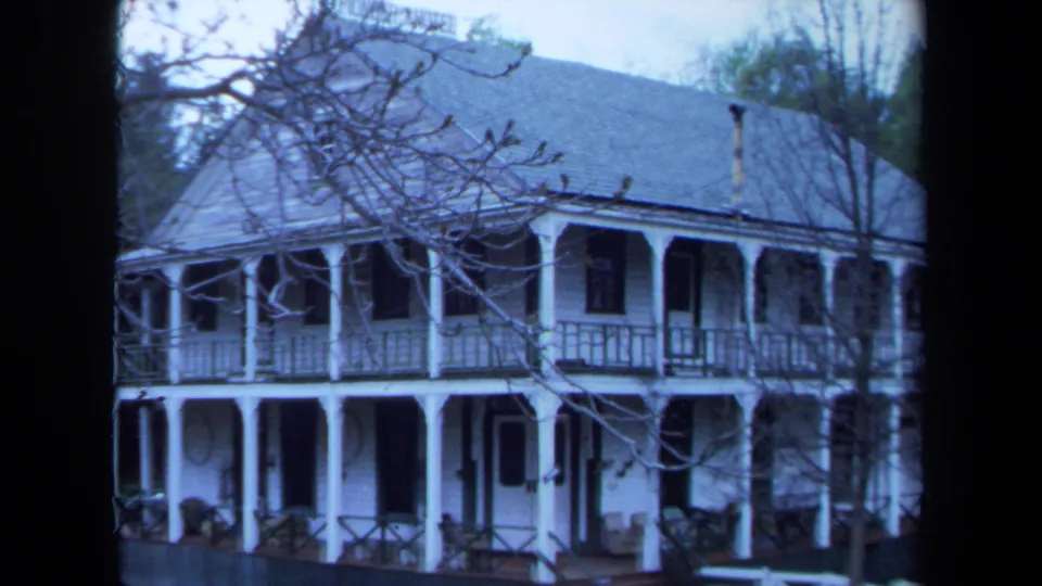 1971 Maine 8mm frame showing a white two-story house with porches, columns, bare trees, and a wooded hillside behind it.
