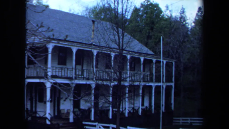 1971 Maine 8mm frame showing the side of a white house with two levels of porch, a flagpole, bare branches, and forest behind.