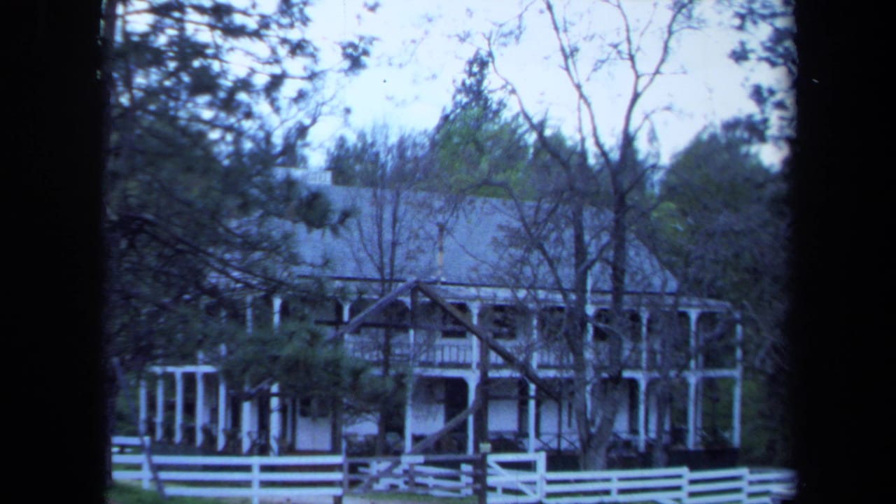 1971 Maine 8mm frame of a large white farmhouse with wraparound porch, trees, and a white picket fence.