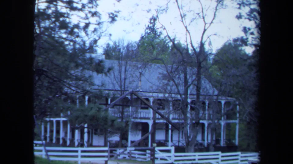 1971 Maine 8mm frame showing the farmhouse exterior through trees, with porch columns and white fencing in front.