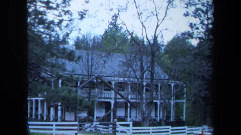 1971 Maine 8mm frame of the same farmhouse seen through trees, with a white fence and shaded porch area.