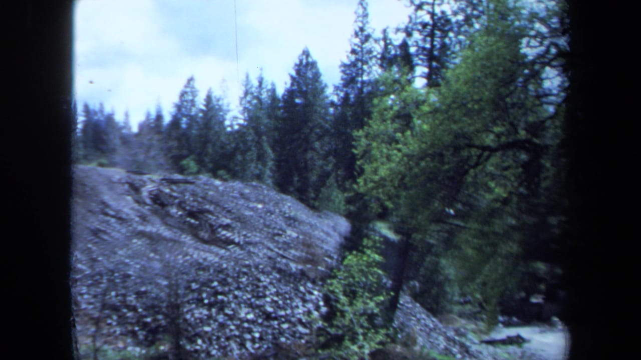 1971 Maine home-movie frame showing a rocky wooded slope with a pale road or clearing at the right.