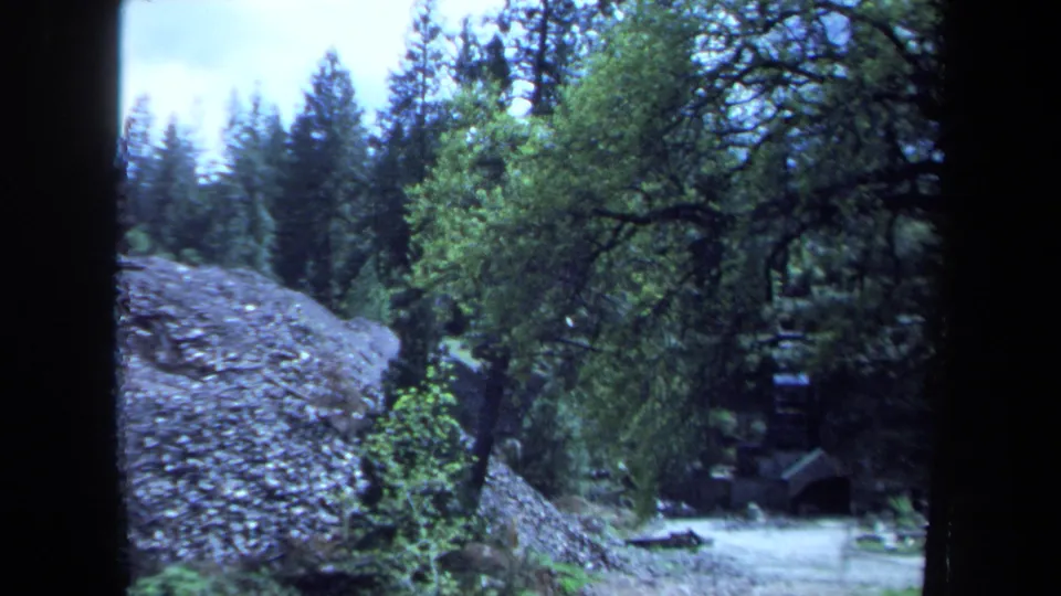 1971 Maine 8mm frame with a large rock face, surrounding trees, and a partially obscured wooden structure below.