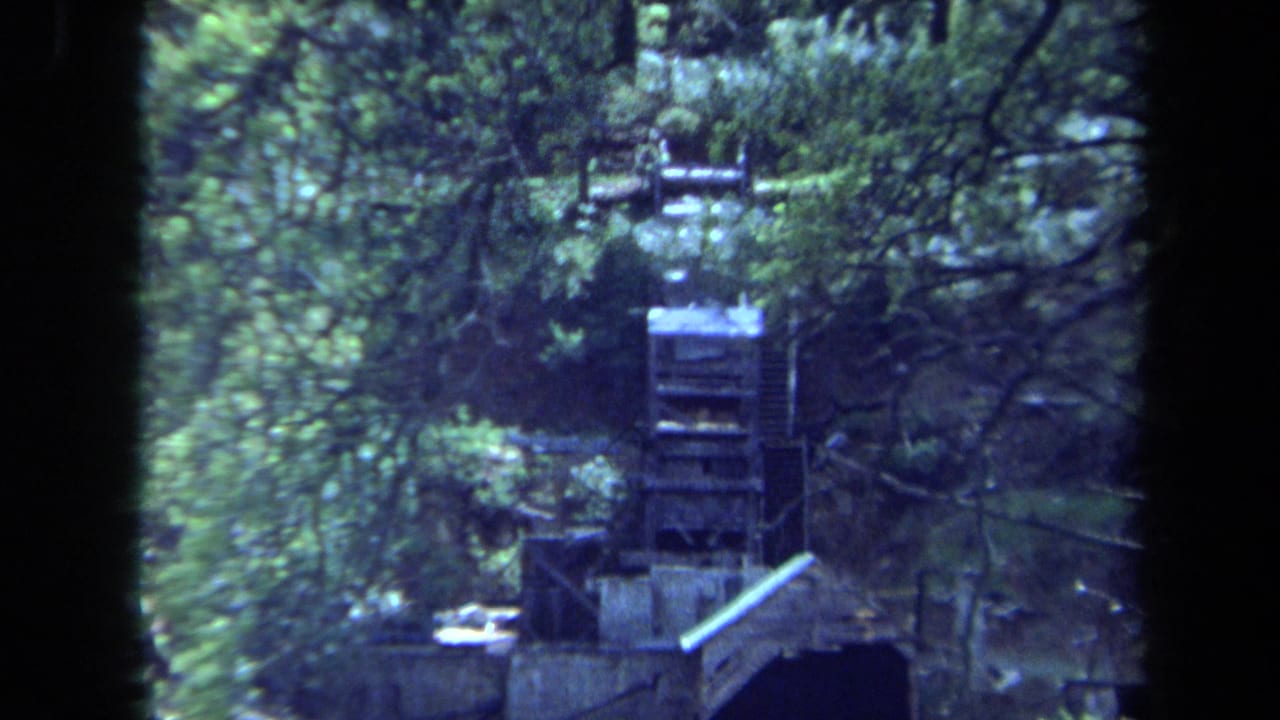 1971 Maine 8mm frame of a wooden waterwheel seen through dense green trees near a stream or mill structure.