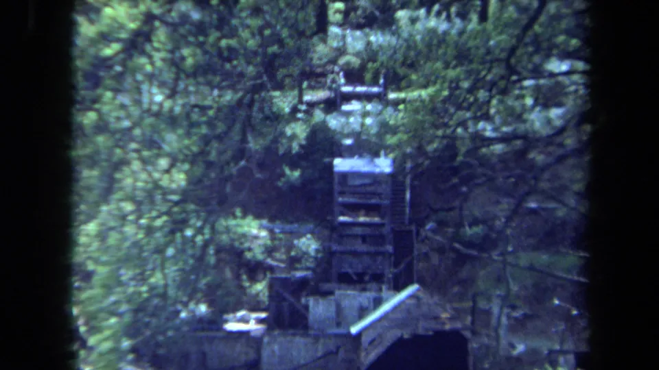 1971 Maine 8mm frame of a wooden waterwheel seen through dense green trees near a stream or mill structure.
