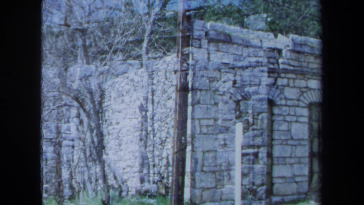Stone corner wall with arched window, trees, and brush in the background.