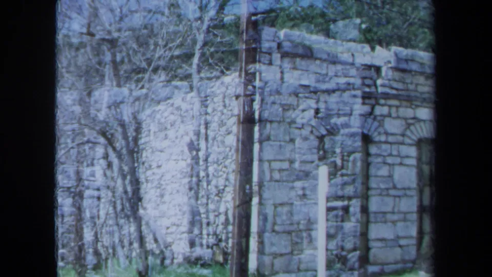 1971 Maine 8mm frame of a weathered stone building corner with an arched window, bare trees, and overgrown countryside brush.