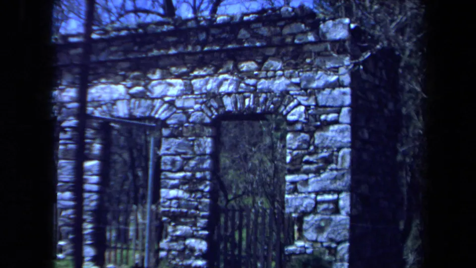 1971 Maine 8mm frame showing an arched stone doorway with metal bars, rough masonry, bare branches, and greenery beyond.