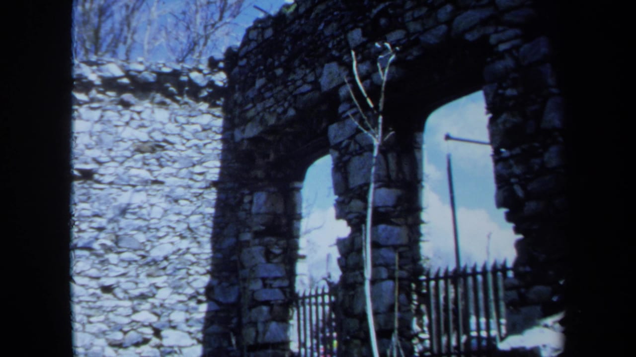 1971 Maine 8mm frame of a crumbling stone wall, arched opening, thin sapling, and black fence in daylight.