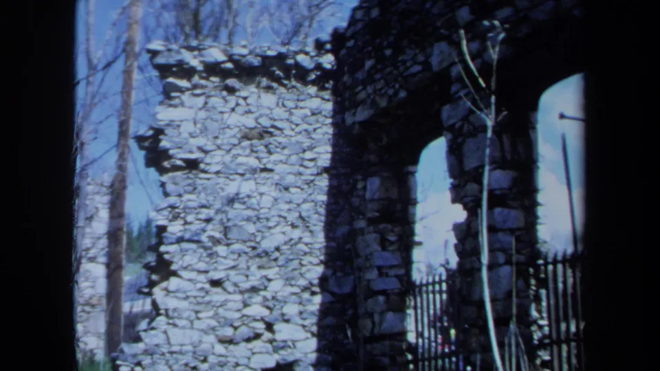1971 Maine 8mm frame showing a stone ruin wall, arched opening, black fence, and leafless trees beyond.