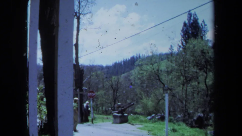 1971 Maine home-movie frame showing a rural roadside, wooded hills, overhead cable, stop sign, and two distant walkers.
