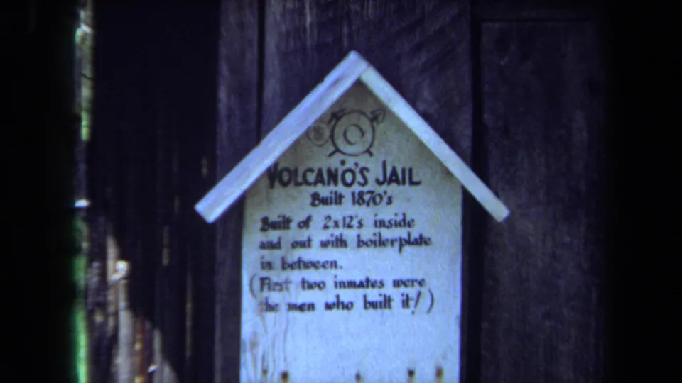 Close 1971 Maine frame of the Volcano's Jail sign, with hand-painted text about its 1870s construction on a dark wooden wall.