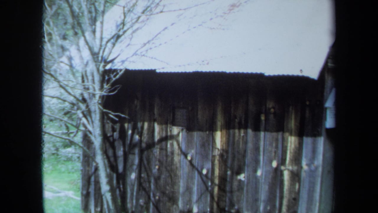1971 Maine home-movie frame of a weathered wooden outbuilding seen through window bars, with bare trees and pale sky nearby.