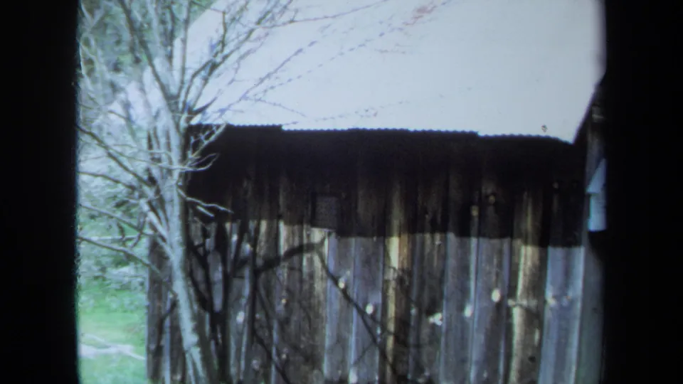 1971 Maine home-movie frame of a weathered wooden outbuilding seen through window bars, with bare trees and pale sky nearby.