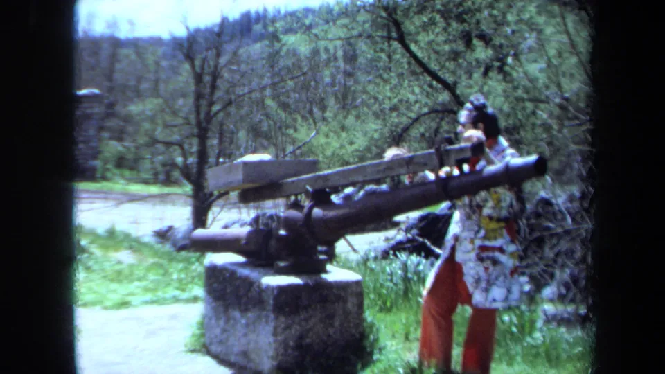 1971 Maine 8mm frame showing a visitor in patterned clothing and sunglasses beside a stationary artillery piece in a wooded setting.