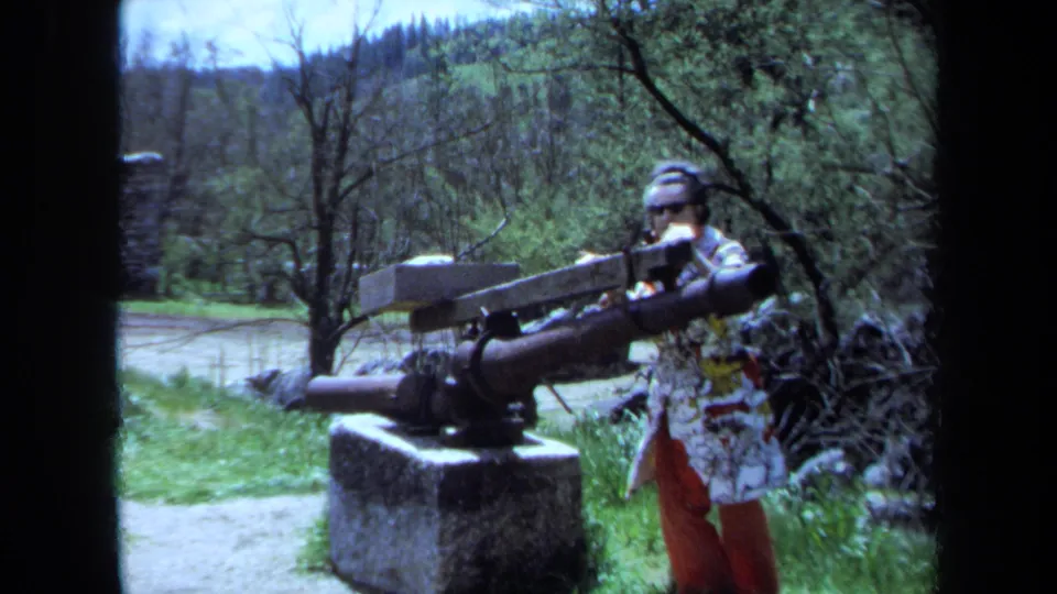 1971 Maine home-movie frame of a visitor by a mounted artillery piece on a stone base, with trees, grass, and low hills in the background.