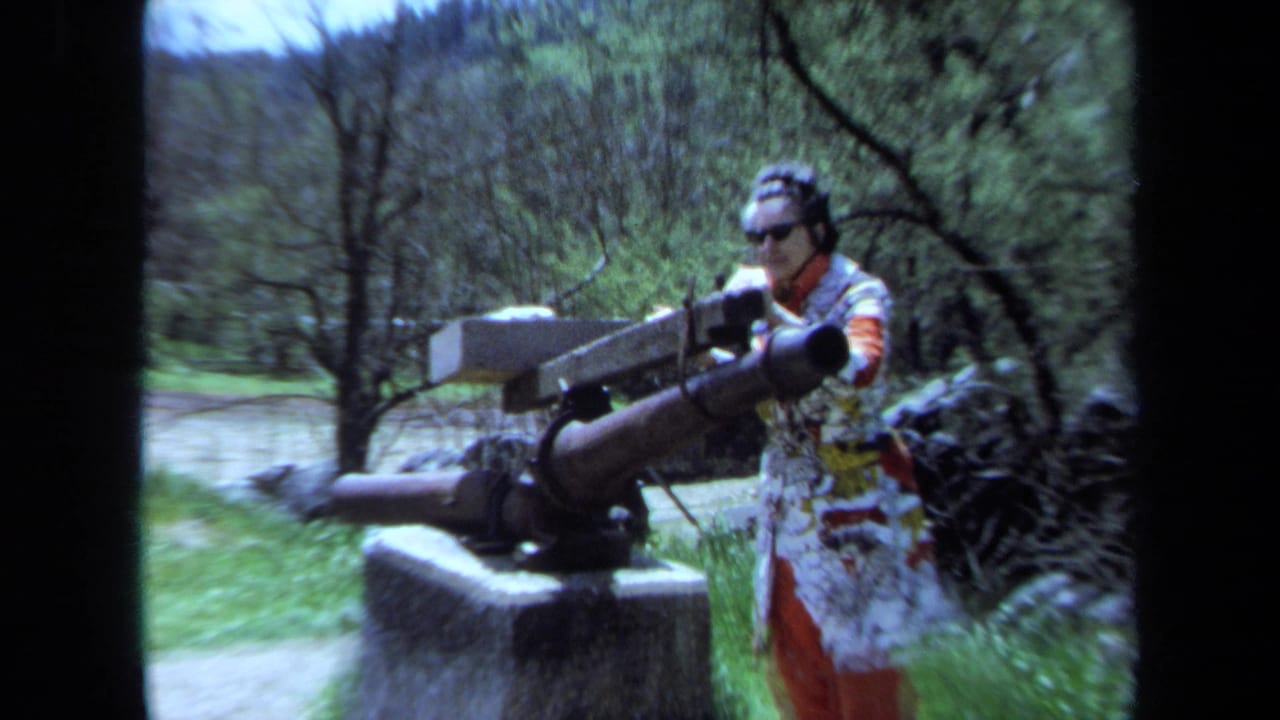 In Maine 1971 home-movie footage, a woman in a patterned jacket stands near a large cannon on a stone base among trees.