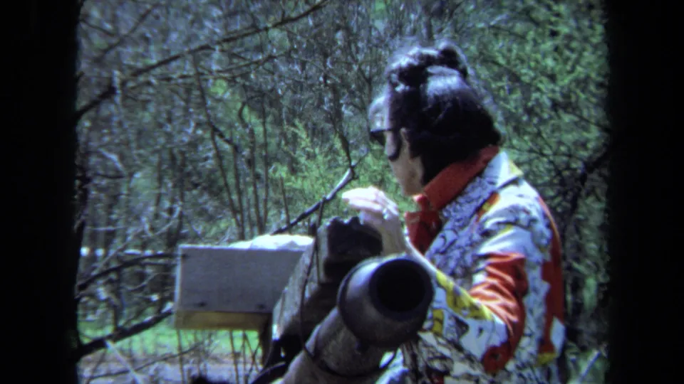 The woman appears to operate the rear mechanism of the cannon, with trees and a small mounted box visible in the background.