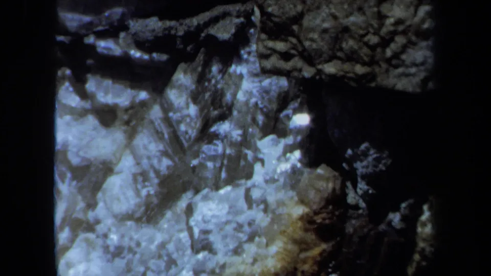 Handheld 1971 Maine frame of jagged stone, pale mineral patches, and a dark crevice under low light.